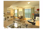 Open-plan interior showing a kitchen sink in the foreground and a furnished living and dining area with a sofa, dining table, and large window with curtains.