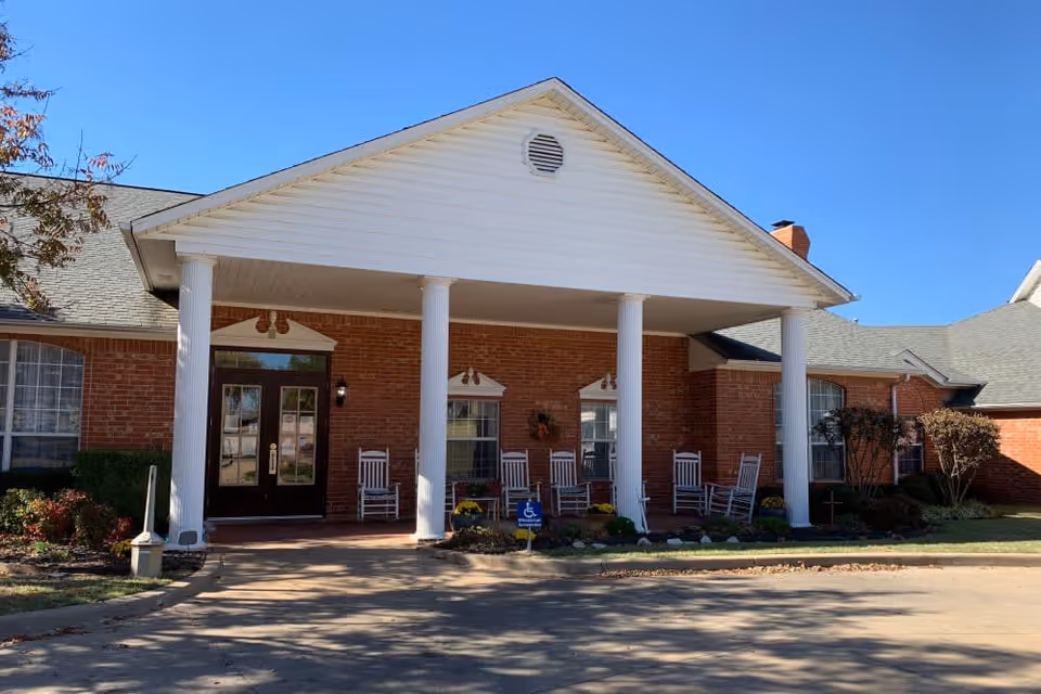 Front exterior view of a brick building with a covered porch supported by four white columns. The porch has several white rocking chairs and a wheelchair accessible sign near the entrance. The sky is clear and blue.