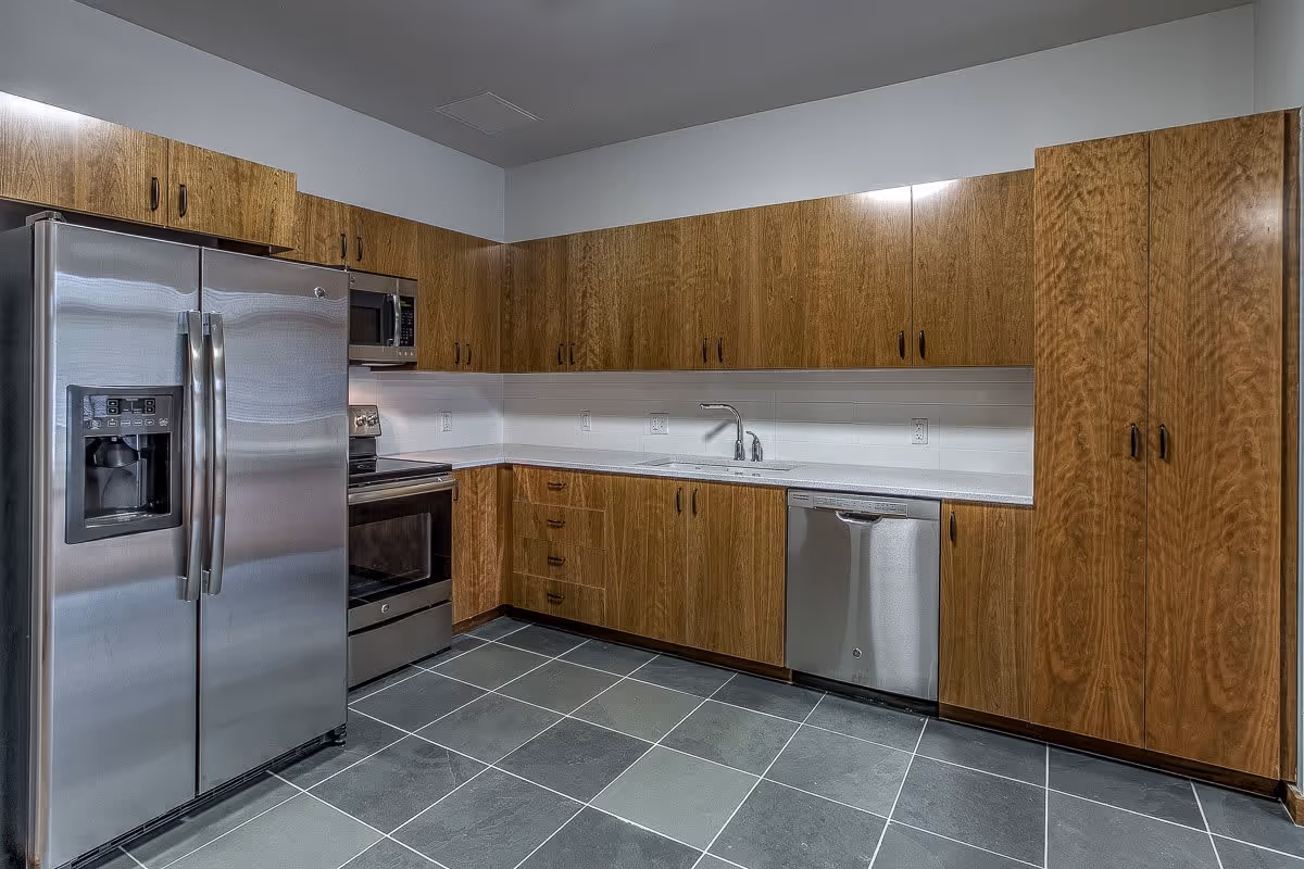 Modern kitchen with stainless steel appliances including a refrigerator, oven, microwave, and dishwasher. The kitchen features wooden cabinets, a white countertop, a sink with a faucet, and gray tiled flooring.