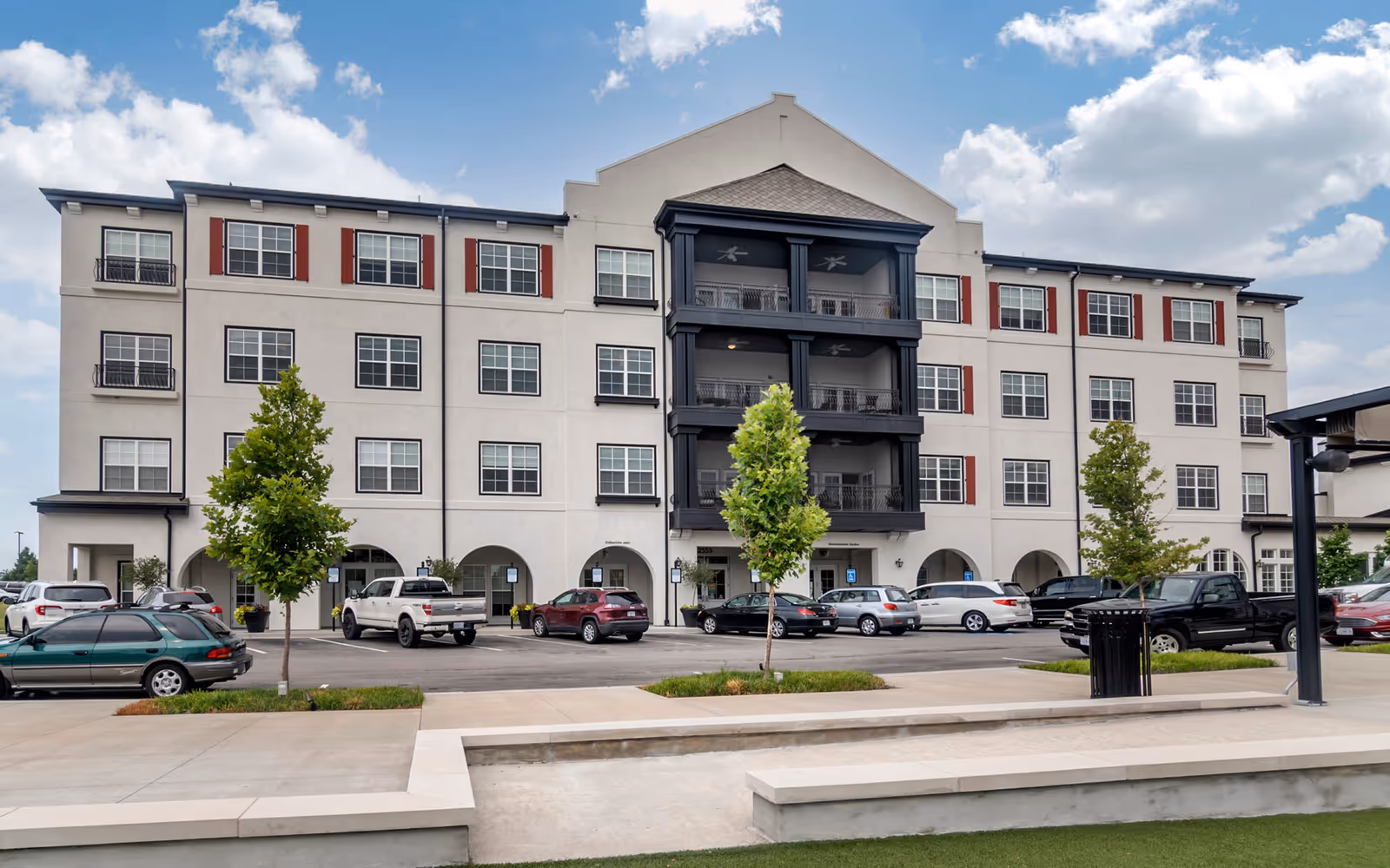 Exterior view of a four-story senior living facility building with multiple windows and balconies. There are several parked cars in front of the building, small trees, and a clear sky with some clouds.