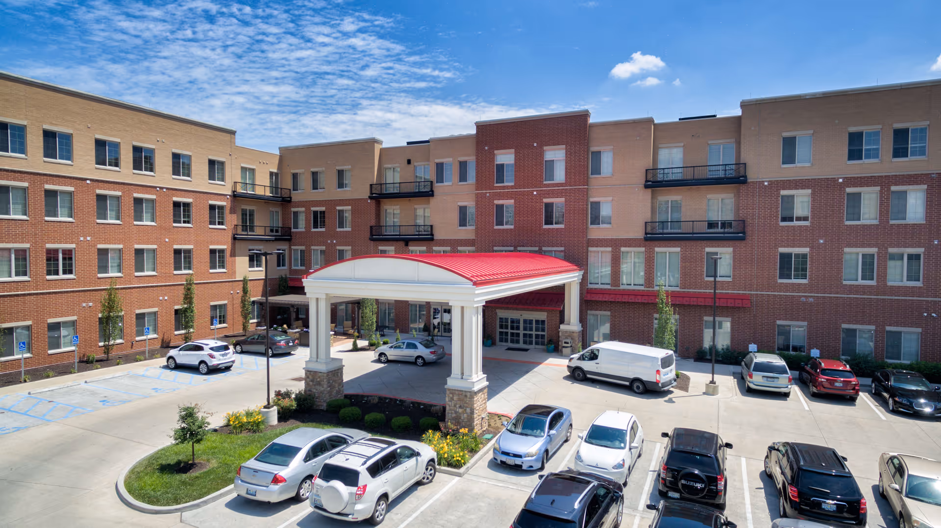 Exterior view of a multi-story assisted living and memory care facility with a covered entrance featuring a red roof. Several cars are parked in the parking lot in front of the building under a partly cloudy blue sky.