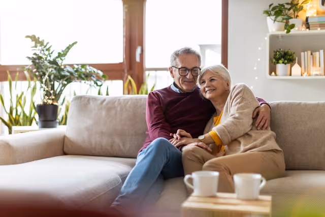 An elderly couple sitting closely together on a beige sofa in a cozy living room, smiling and holding hands. The room has large windows with plants on the windowsill and shelves with decorative items in the background.