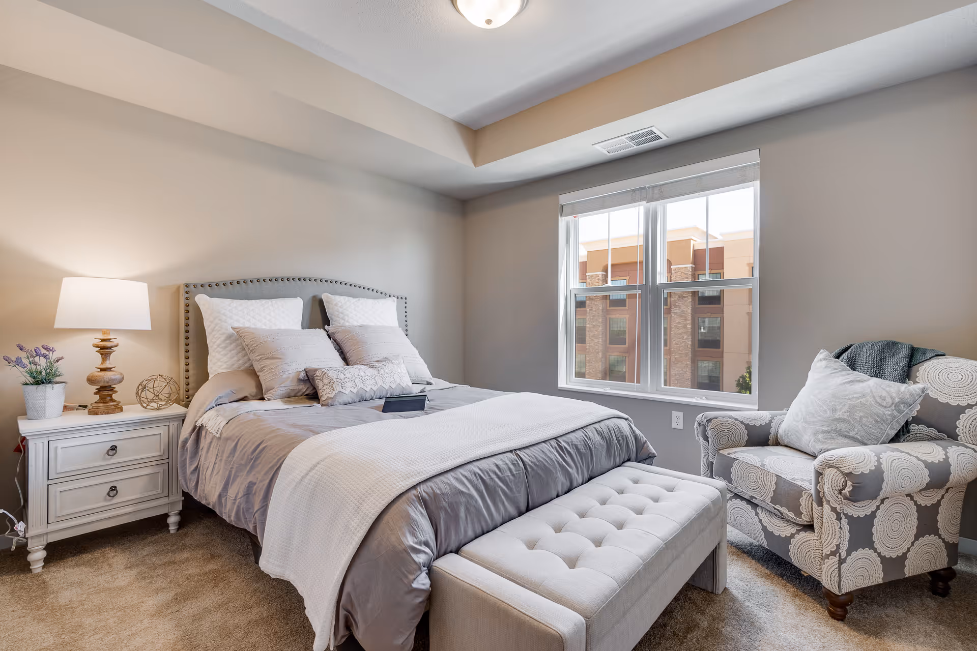 Bright, neatly staged bedroom with a bed, nightstand and lamp, window, upholstered bench and patterned armchair.
