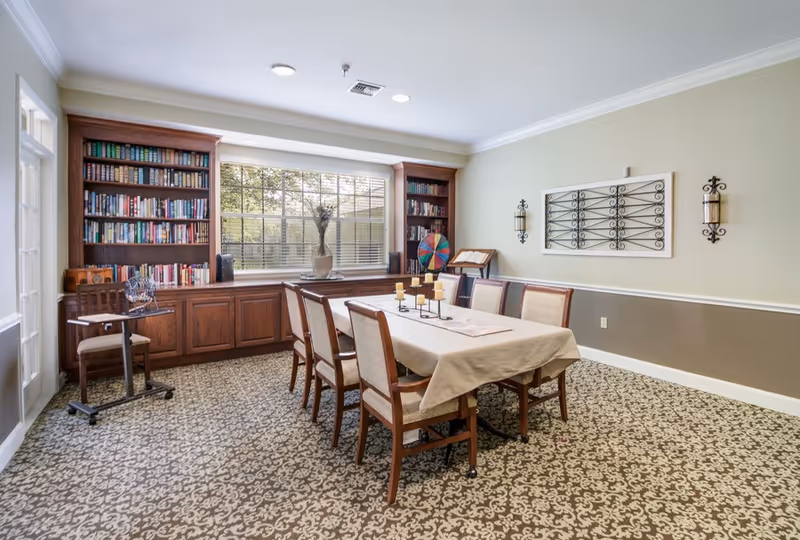 A bright communal dining room with a long table and chairs, built-in bookshelves under a large window, and decorative wall accents.