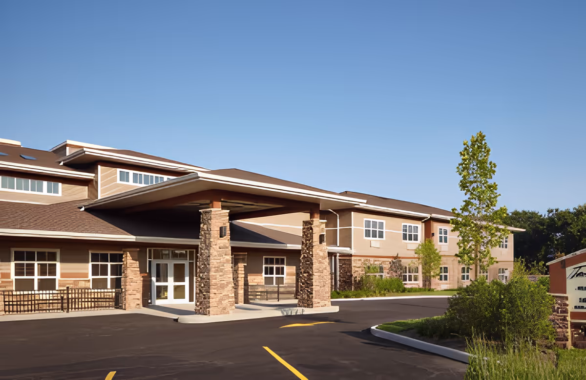 Entrance and facade of a two-story senior living facility with a covered porte-cochère, stone columns, and a landscaped driveway under a clear blue sky.