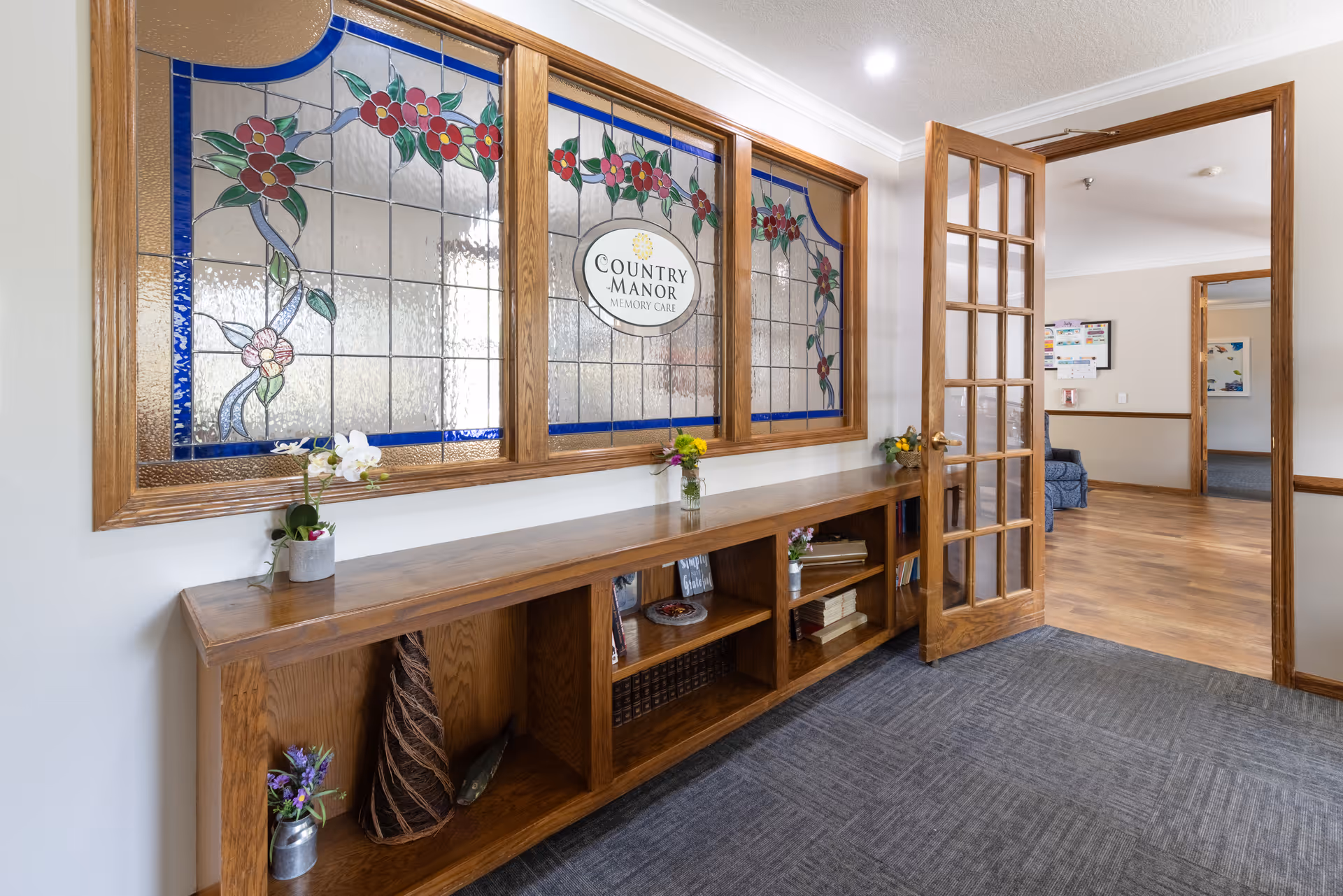 Interior view of a senior living facility hallway with a wooden bookshelf holding decorative items and flowers. Above the bookshelf is a large stained glass window featuring floral designs and the text 'Country Manor Memory Care'. A wooden framed glass door is open, leading to another room with wood flooring and a bulletin board on the wall.