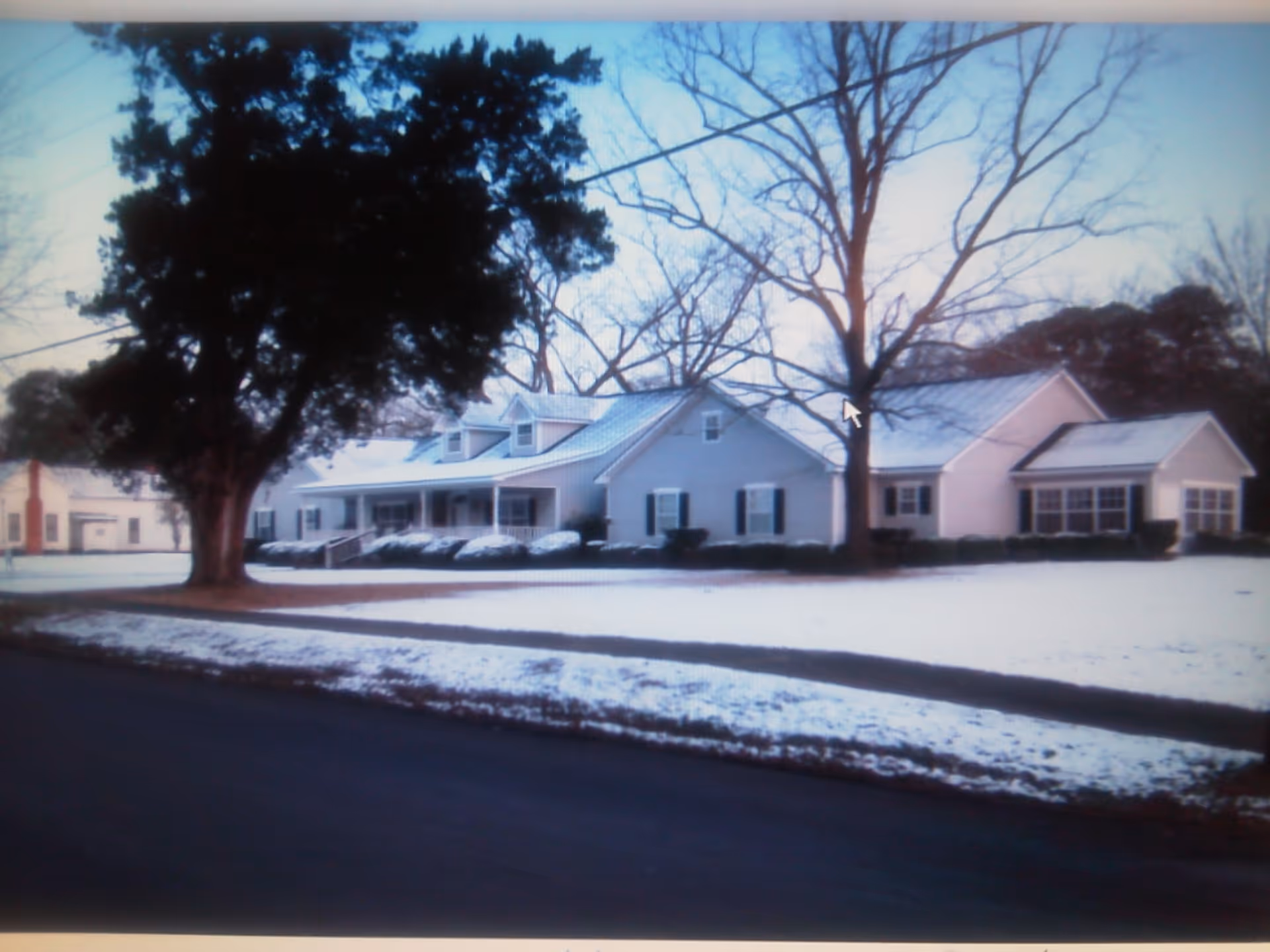 One-story light-colored building with a covered front porch and large trees, set behind a snow-covered lawn and road.