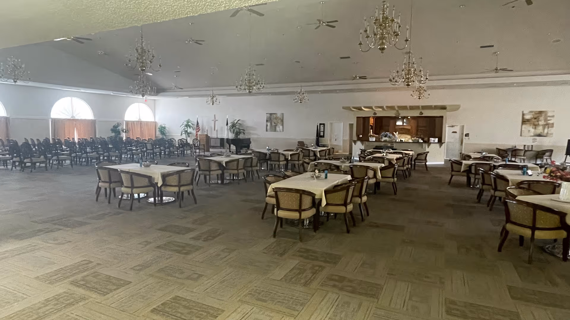 Large dining room with multiple round tables covered with beige tablecloths and surrounded by cushioned chairs. The room has chandeliers hanging from the ceiling, large arched windows with curtains, and a small stage area with a piano and flags in the background. There is also a kitchen or serving area visible at the back of the room.