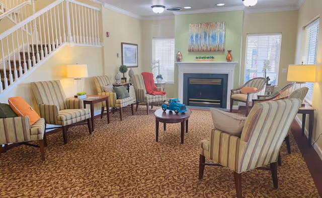 A cozy living room area in a senior living facility featuring multiple striped armchairs arranged around a small round wooden table with a blue toy truck on it. The room has a patterned carpet, a fireplace with decorative items on the mantel, two windows with blinds, and soft lighting from table lamps and ceiling fixtures. A staircase with white railings is visible on the left side.