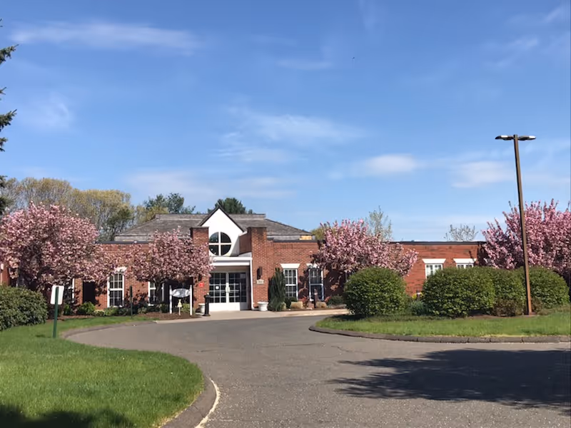 Front exterior view of a single-story brick building with a peaked roof entrance, surrounded by blooming pink trees and green bushes under a blue sky.