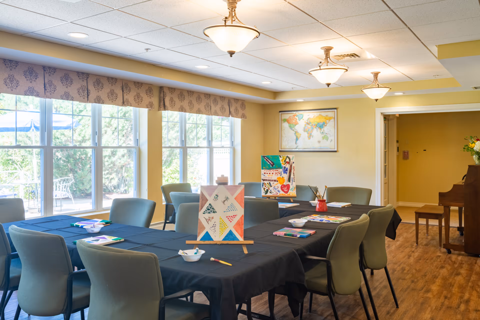 A bright activity room with large windows letting in natural light. There are two tables covered with black tablecloths, surrounded by green cushioned chairs. On the tables are art supplies, including paintbrushes, bowls, and colorful artwork on small easels. A world map hangs on the wall, and a piano is visible in the corner of the room. The room has a warm, inviting atmosphere with wooden flooring and ceiling lights.