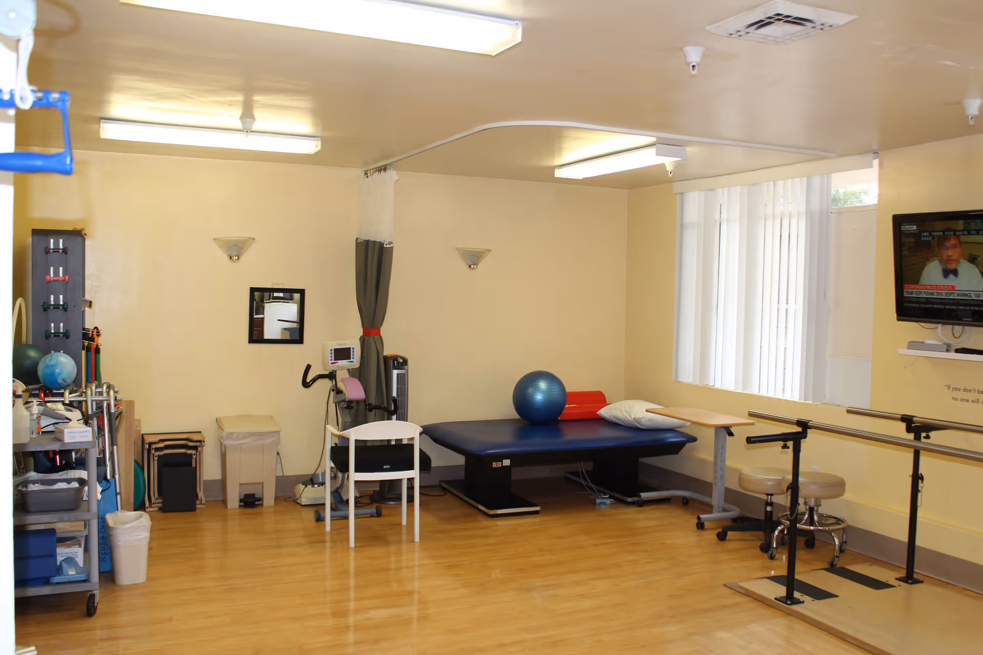 A physical therapy or rehabilitation room with wooden flooring, a blue therapy table with a pillow and exercise ball on it, parallel bars for walking exercises, a stationary exercise bike, a white chair, and various exercise equipment and supplies on shelves. The room has beige walls, fluorescent ceiling lights, a wall-mounted TV, and a window with vertical blinds.