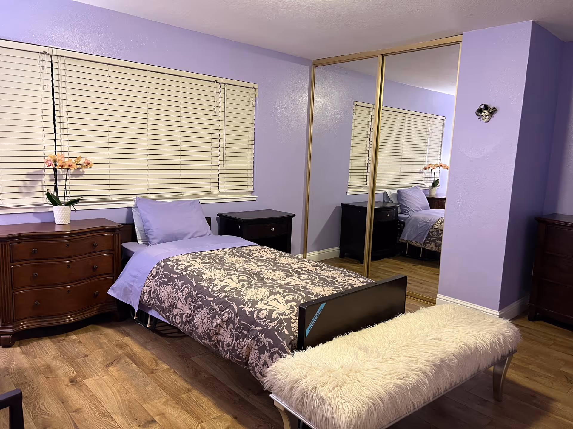A bedroom with lavender walls and wooden flooring featuring a single bed with a patterned gray and white comforter and lavender pillow and sheet. There is a wooden dresser with a potted orchid on top, a small dark nightstand, and a mirrored closet door reflecting the room. A fluffy white bench is placed at the foot of the bed.