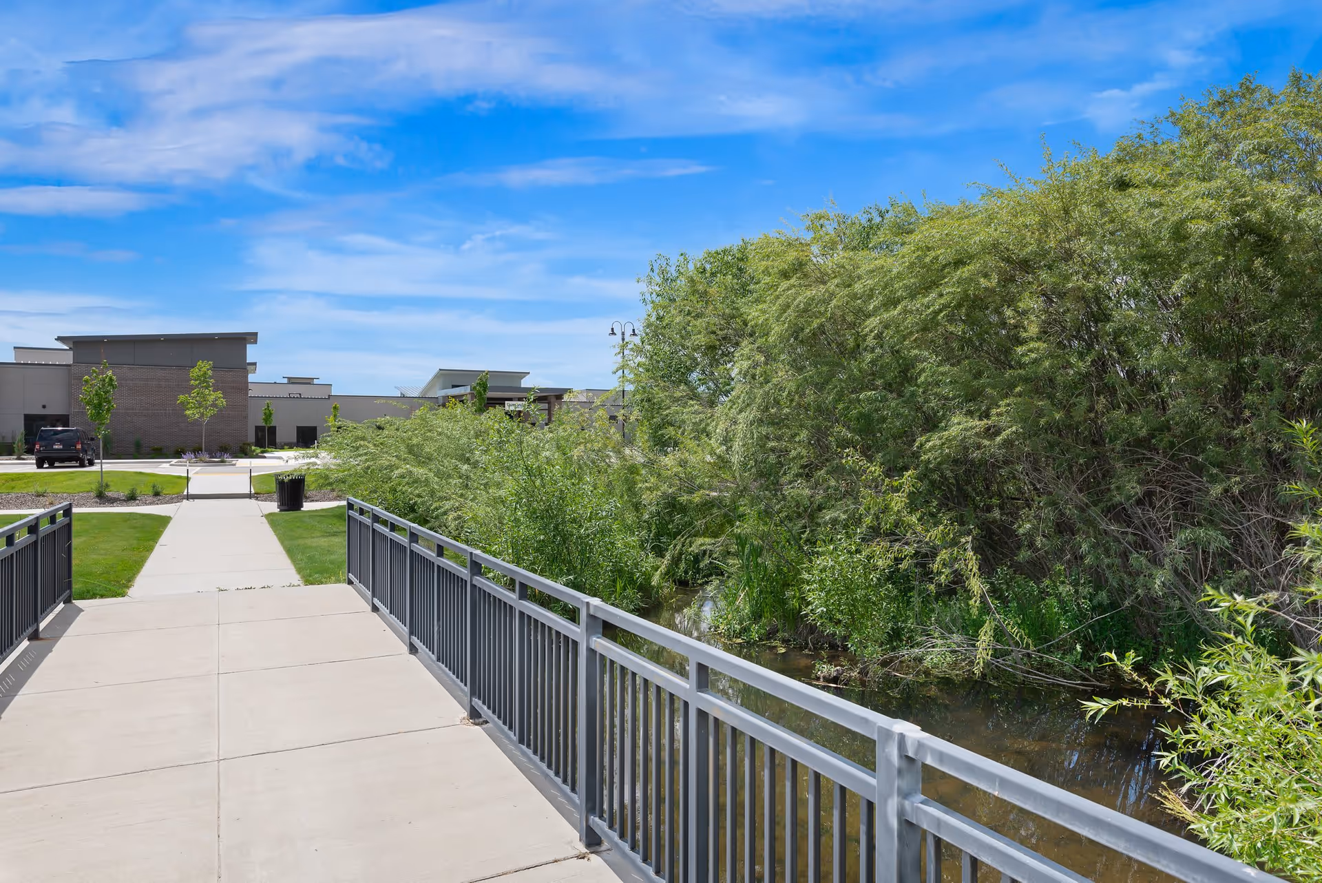A paved walkway and metal-railed bridge over a small waterway leading toward a modern senior living building with trees and shrubs on a sunny day.