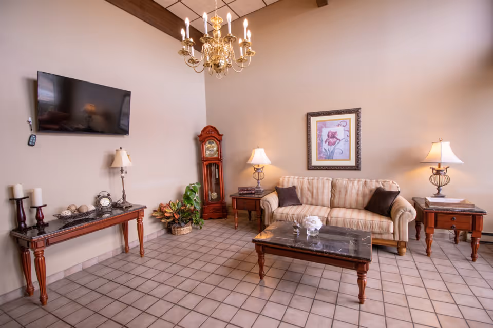 A cozy living room with a beige striped sofa adorned with two dark pillows, a dark marble coffee table, two wooden side tables each with a lamp, a tall wooden grandfather clock, a wall-mounted flat-screen TV, a decorative console table with candles and a clock, a basket with plants, and a framed floral artwork on the wall. The room has tiled flooring and a brass chandelier hanging from a ceiling with wooden beams.