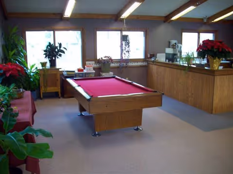 A communal room with a red-felt pool table, wood-paneled reception counter, windows, and potted plants.