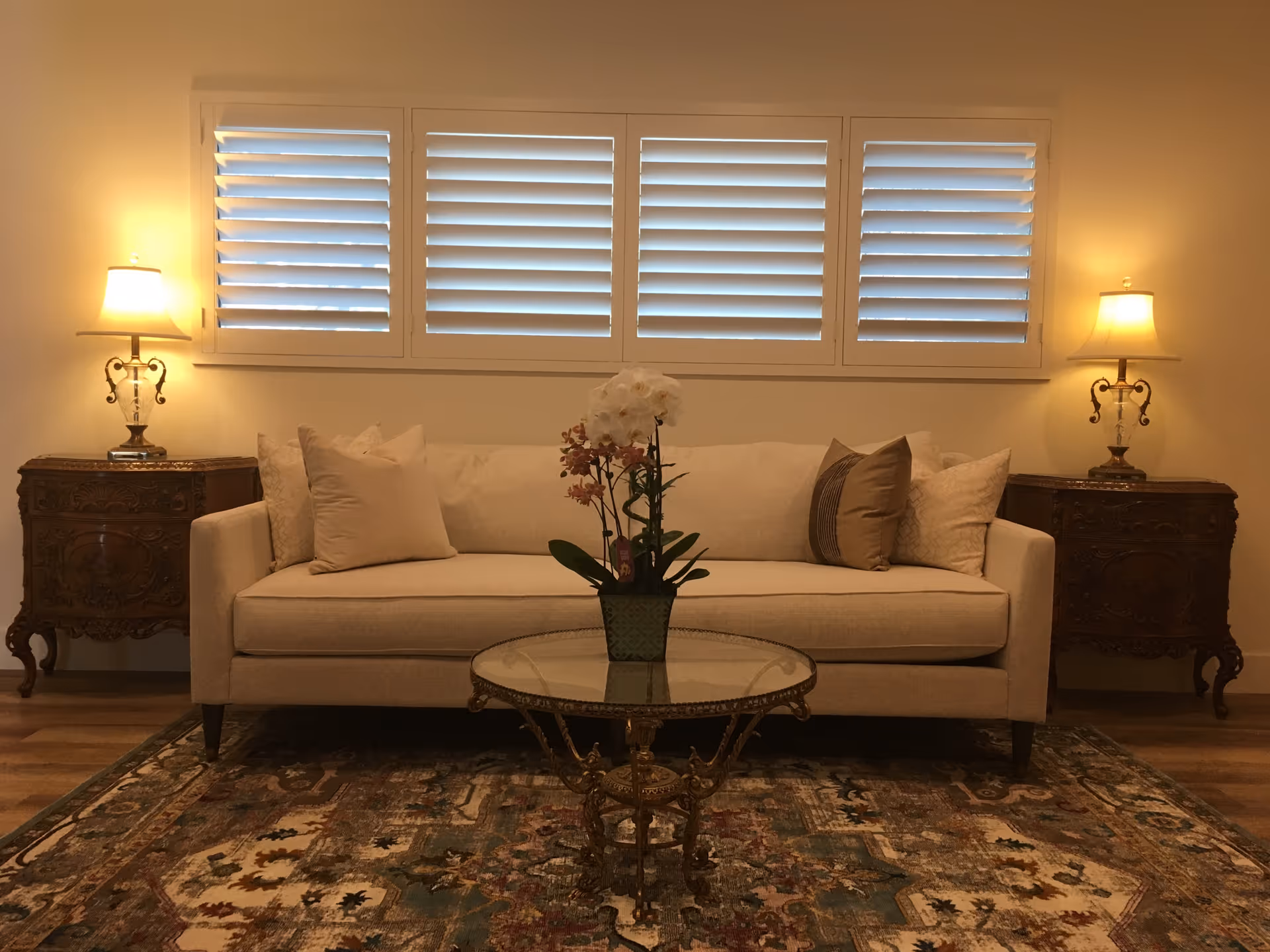 Cream sofa with pillows flanked by two ornate side tables and lamps, a glass coffee table with an orchid, and shuttered windows above.