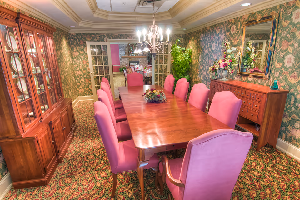 Formal dining room with a long wooden table surrounded by pink upholstered chairs, a china cabinet, floral wallpaper, and a chandelier.