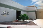 Exterior view of Legacy Rehab & Care Center building with a small landscaped area featuring a palm plant and rocks under a partly cloudy sky.