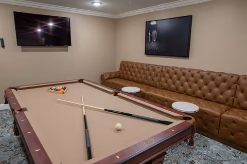 A recreational room with a beige pool table in the center, two pool cues and a white cue ball on the table, a brown tufted leather sofa along the wall with two small round white tables in front of it, a flat-screen TV mounted on the wall, and a framed black and white picture hanging above the sofa.