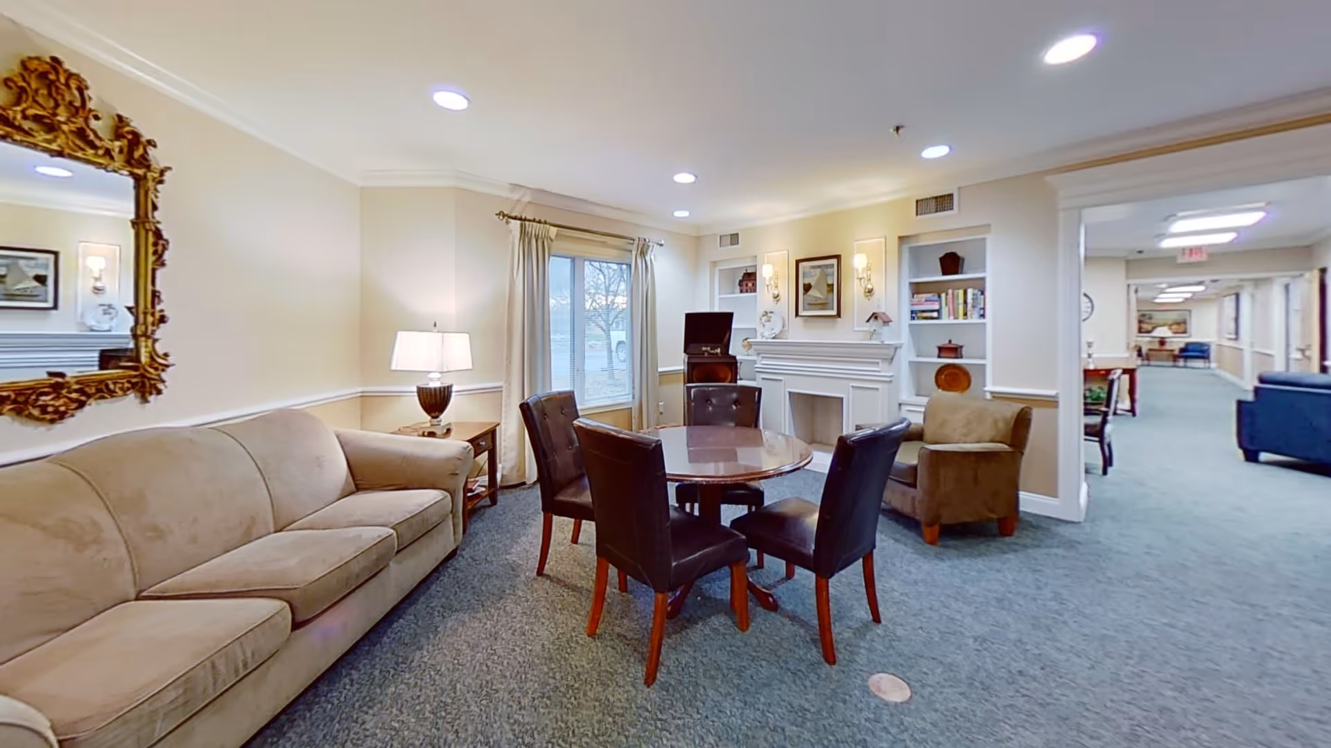A cozy common area in a senior living facility featuring a beige sofa, a round wooden table with four dark leather chairs, a brown armchair, a side table with a lamp, a decorative fireplace with built-in shelves, and a large ornate mirror on the wall. The room is well-lit with recessed ceiling lights and has a window with curtains overlooking the outside.