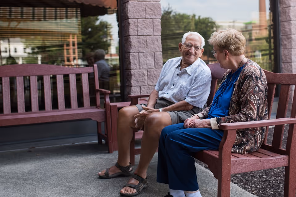 An elderly man and woman sitting on wooden benches outside a building, engaged in conversation and smiling. The man is wearing a light-colored short-sleeve shirt and shorts, while the woman is dressed in a blue outfit with a patterned cardigan. The background shows a window reflecting outdoor scenery.