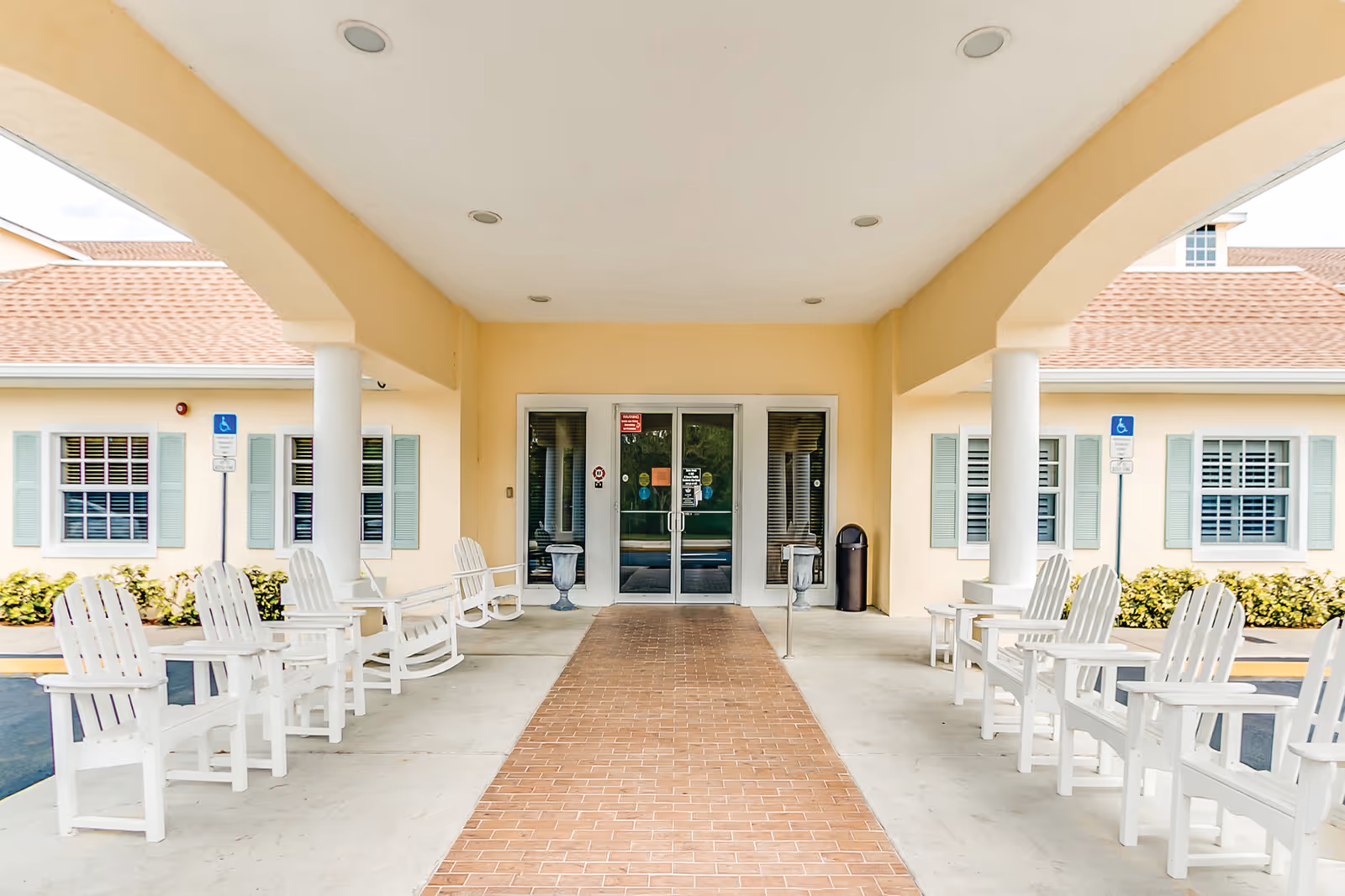 Covered entrance area of Cedar Creek Assisted Living with white rocking chairs lined up on both sides, beige walls, and double glass doors at the center. There are two handicap parking signs visible near the building windows with green shutters.