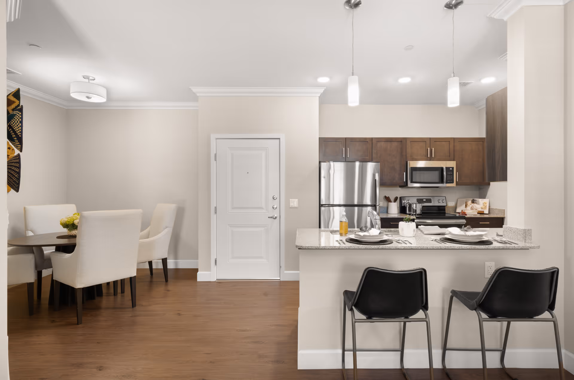 Interior view of a modern kitchen and dining area in a senior living facility. The kitchen features dark wood cabinets, stainless steel refrigerator, microwave, and stove. A granite countertop bar with two black chairs is set with plates and glasses. To the left, there is a round dining table with four beige upholstered chairs and a small flower arrangement. The floor is wooden, and the walls are painted light beige.