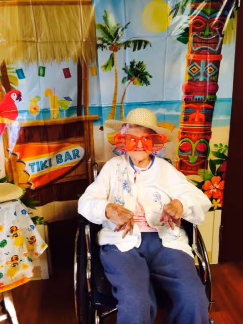 An elderly person sitting in a wheelchair wearing a straw hat and oversized red sunglasses, posing in front of a colorful tropical-themed backdrop featuring a tiki bar, palm trees, a tiki statue, and a beach scene.