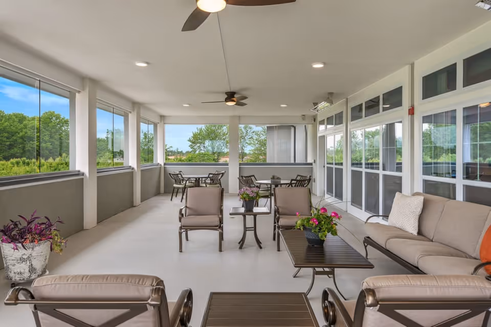 A spacious, covered outdoor patio area with cushioned chairs, sofas, and tables arranged for seating and socializing. The patio is enclosed with large windows offering views of green trees and blue sky outside. Ceiling fans and recessed lighting are installed on the ceiling, and potted plants add a touch of greenery to the space.
