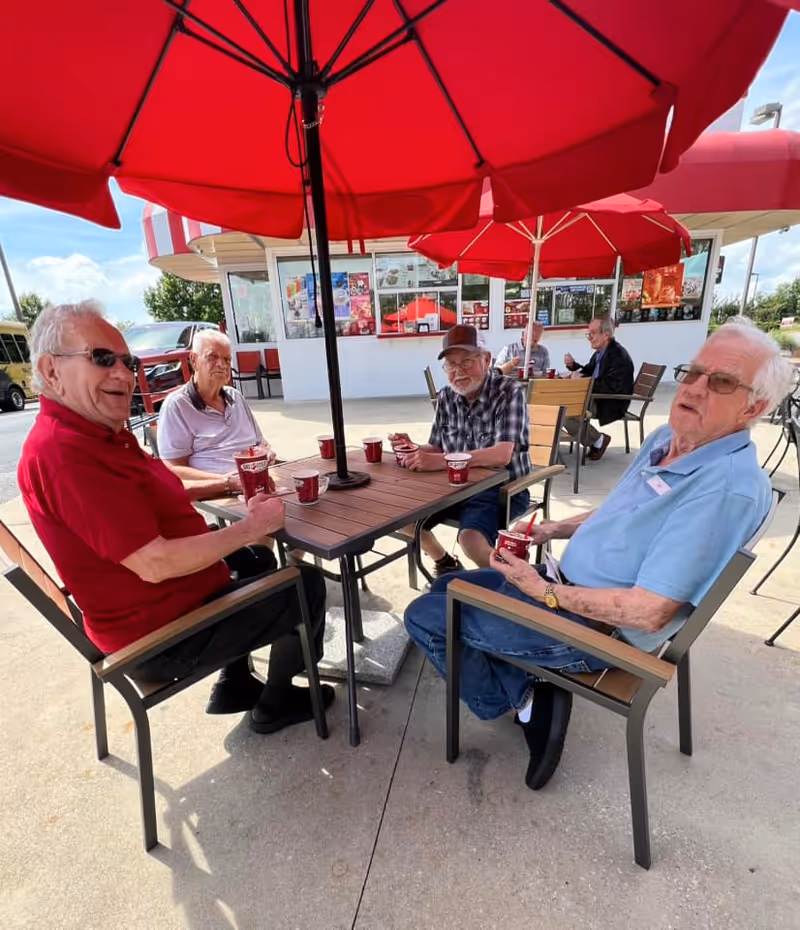 Four elderly men sitting around a wooden table under a large red umbrella outside a building, each holding a cup of ice cream. Two more elderly men are seated at another table in the background. The setting appears to be a casual outdoor dining or social area on a sunny day.