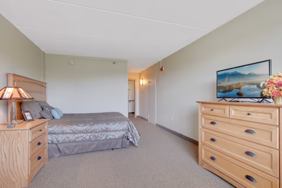 A senior living facility bedroom with a bed covered in a patterned gray bedspread, a wooden nightstand with a lamp and picture frame, a wooden dresser with a flat-screen TV on top, and a vase of flowers. The room has beige walls and carpeted floor.