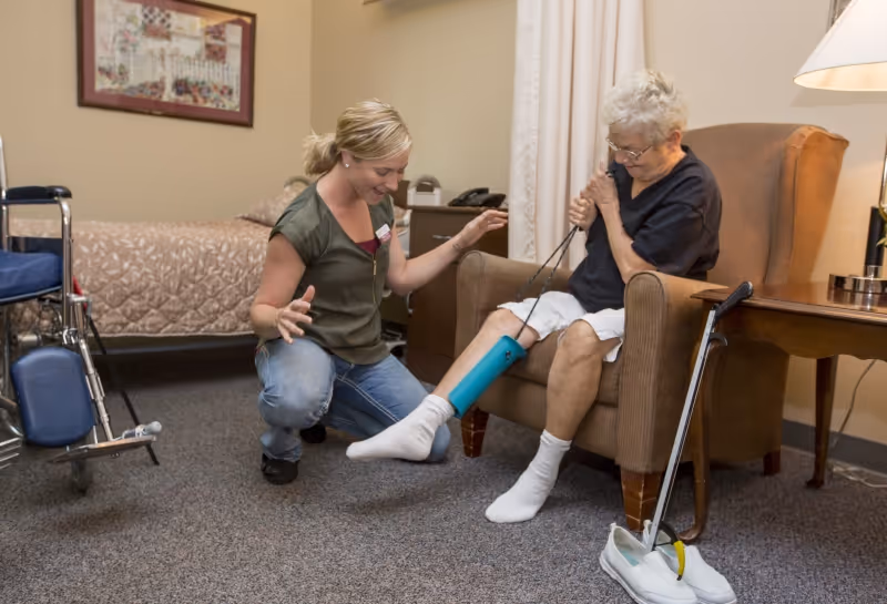 An elderly woman sitting in a brown armchair using a leg exerciser device while a caregiver kneels beside her, smiling and encouraging her. The room has a bed, a wheelchair, a walking cane, a side table with a lamp, and a framed picture on the wall.