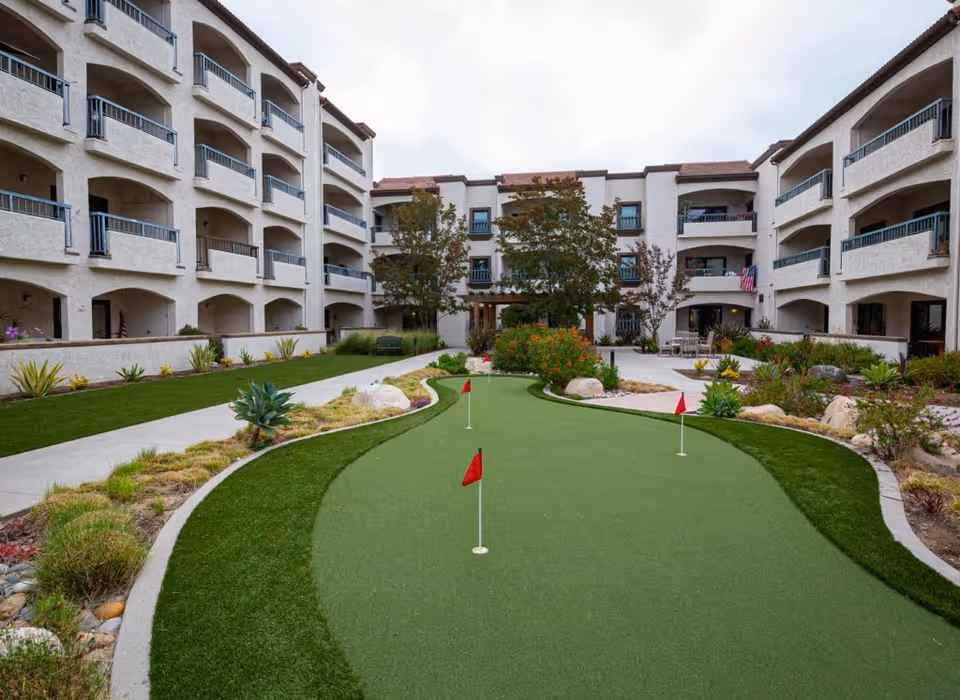 Outdoor courtyard area of Casa de las Campanas featuring a putting green with three red flags, surrounded by landscaped plants and shrubs. The courtyard is enclosed by a multi-story building with balconies and arches.