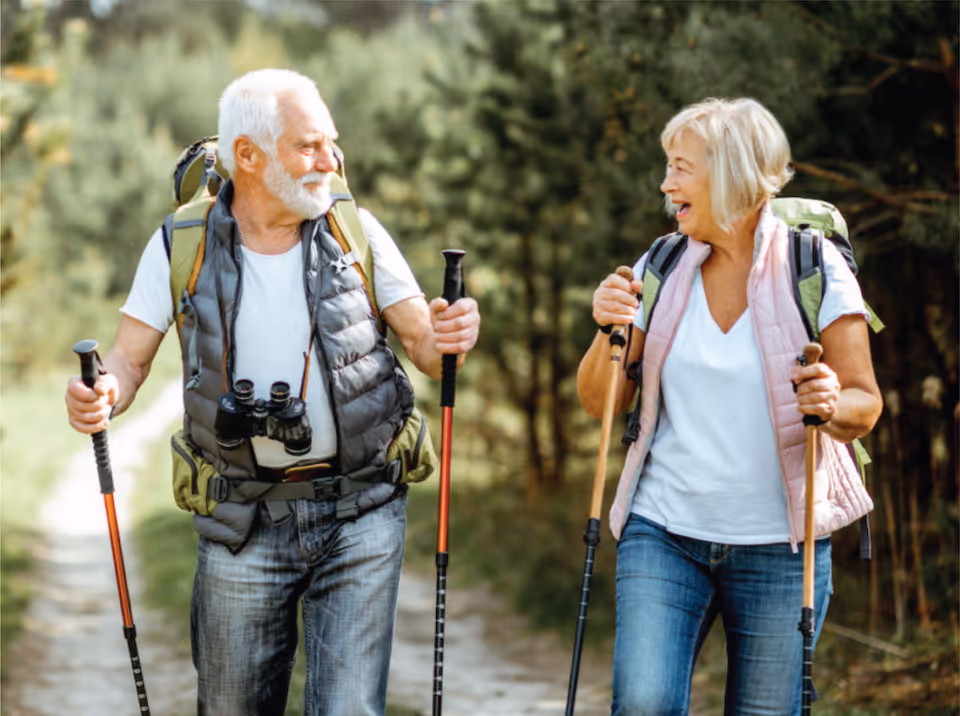 An elderly man and woman hiking on a forest trail, both carrying backpacks and using walking sticks, smiling and looking at each other.