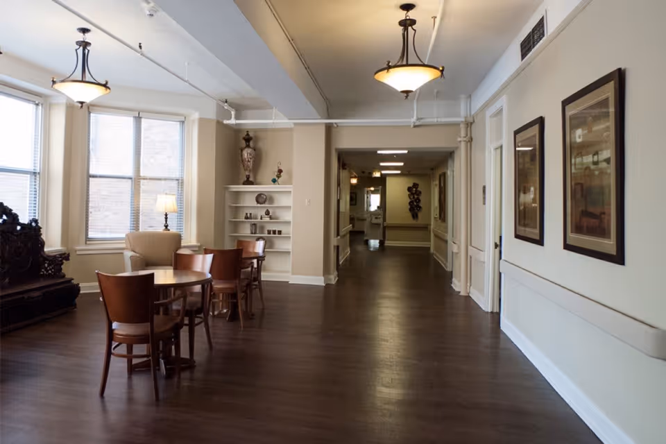 Bright communal lounge with small round tables and chairs, an armchair by large windows, decorative shelving, and a long hallway.