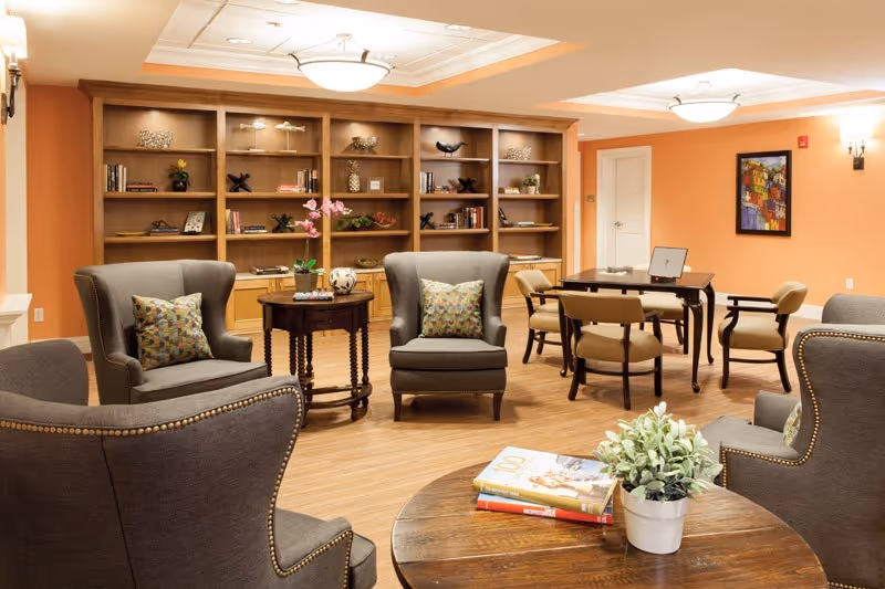 A cozy common area with four gray armchairs arranged around a wooden coffee table that has a small potted plant and books on it. Behind the chairs is a wooden bookshelf filled with decorative items and books. There is also a table with four chairs in the background, a colorful framed painting on the orange wall, and warm ceiling lights illuminating the room.