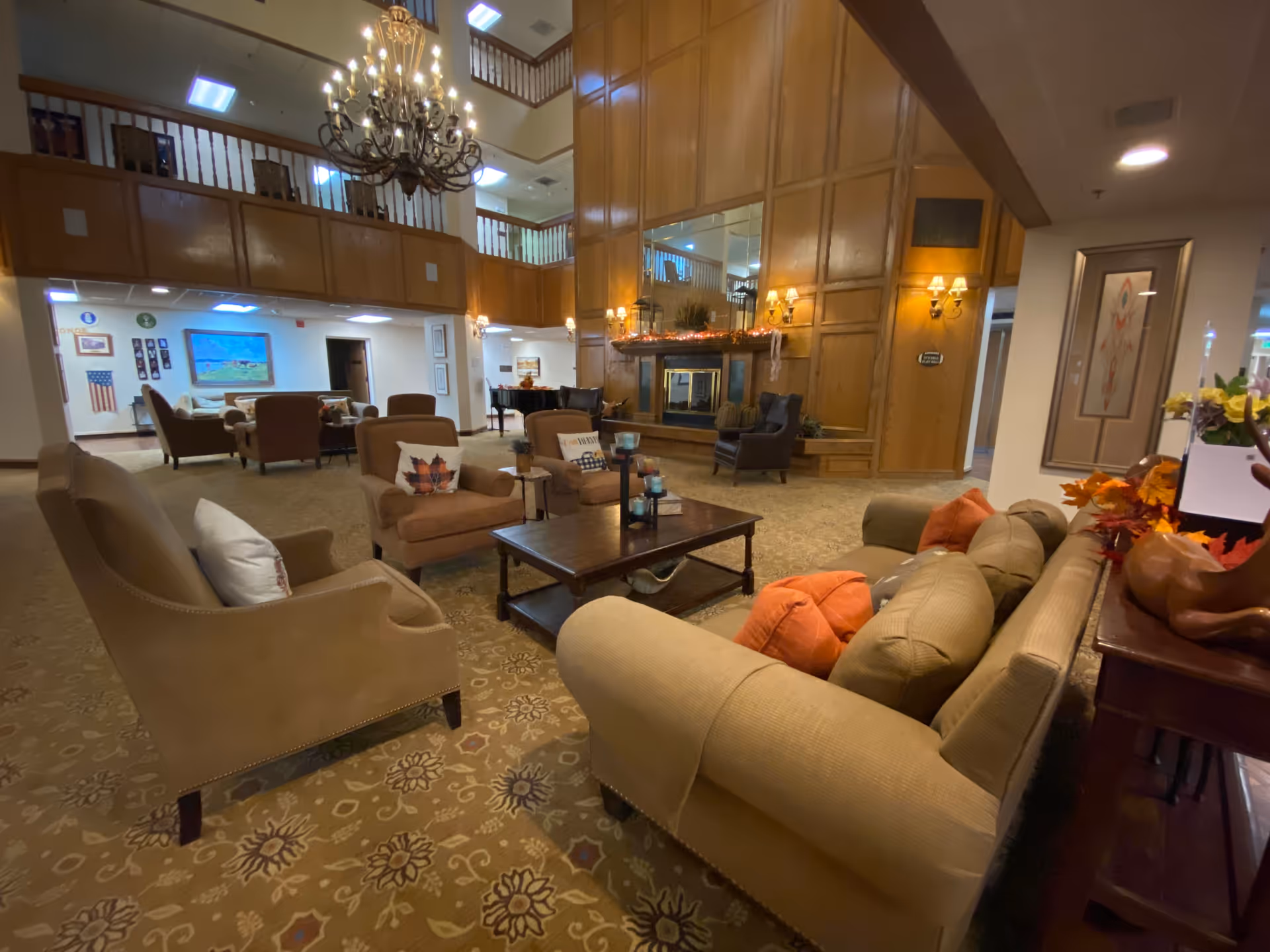 A spacious and warmly decorated living room area in Heritage Place Independent Living featuring multiple comfortable armchairs and sofas arranged around a central wooden coffee table. The room has a high ceiling with a large chandelier, wood-paneled walls with a fireplace, and decorative lighting fixtures. There are autumn-themed decorations on a side table and the fireplace mantel, and a balcony railing is visible on the upper level.