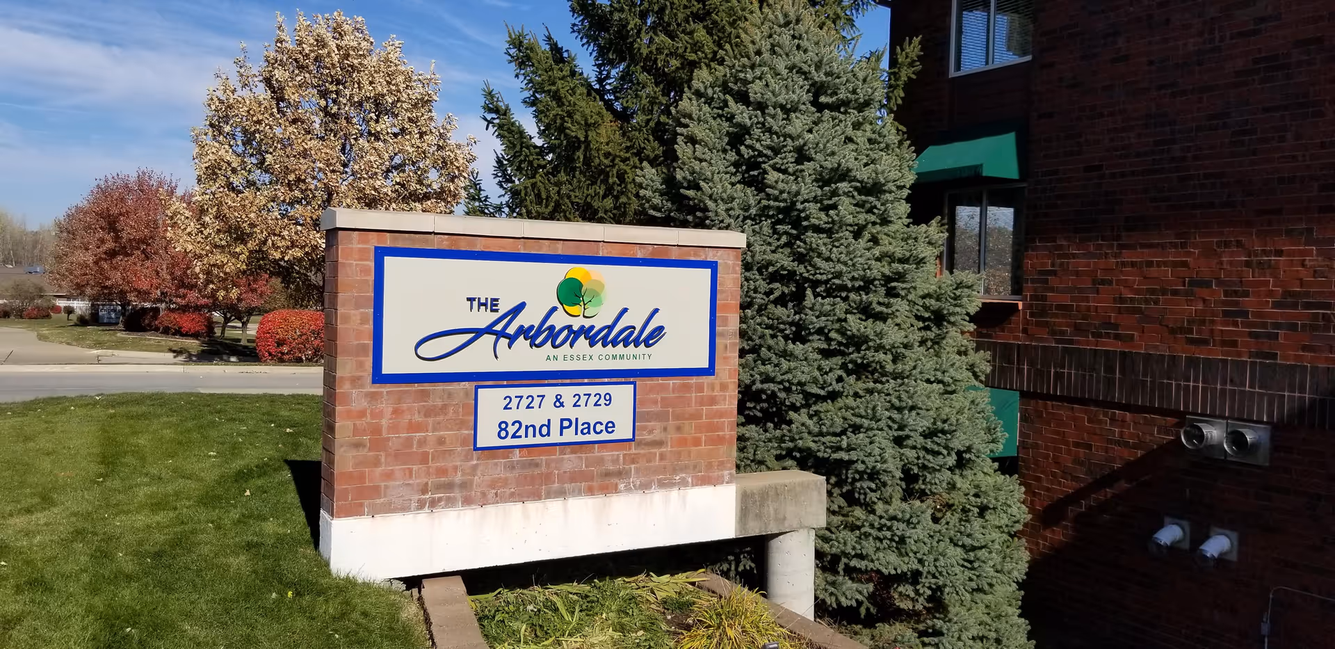 A brick sign outside a building displaying the name 'The Arbordale, An Essex Community' with the address '2727 & 2729 82nd Place'. The sign is surrounded by green grass, trees with autumn foliage, and part of a brick building is visible on the right side.