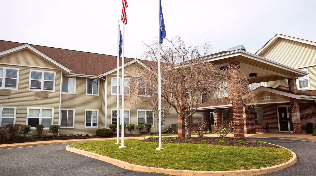 Exterior view of a senior living facility building with beige siding and brick entrance. There are three flagpoles with flags in front of the building, a small landscaped area with grass and a tree, and a driveway leading to the entrance.