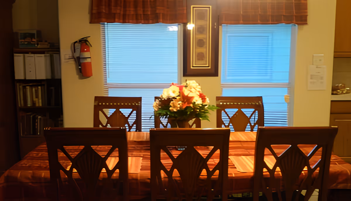 A dining room with a wooden table covered by a brown tablecloth and six matching wooden chairs. A floral centerpiece with white and red flowers is placed in the middle of the table. Behind the table are two windows with closed blinds and brown valances. A fire extinguisher is mounted on the wall to the left, and there is a bookshelf with books and binders. On the right side, part of a kitchen counter and cabinets are visible.