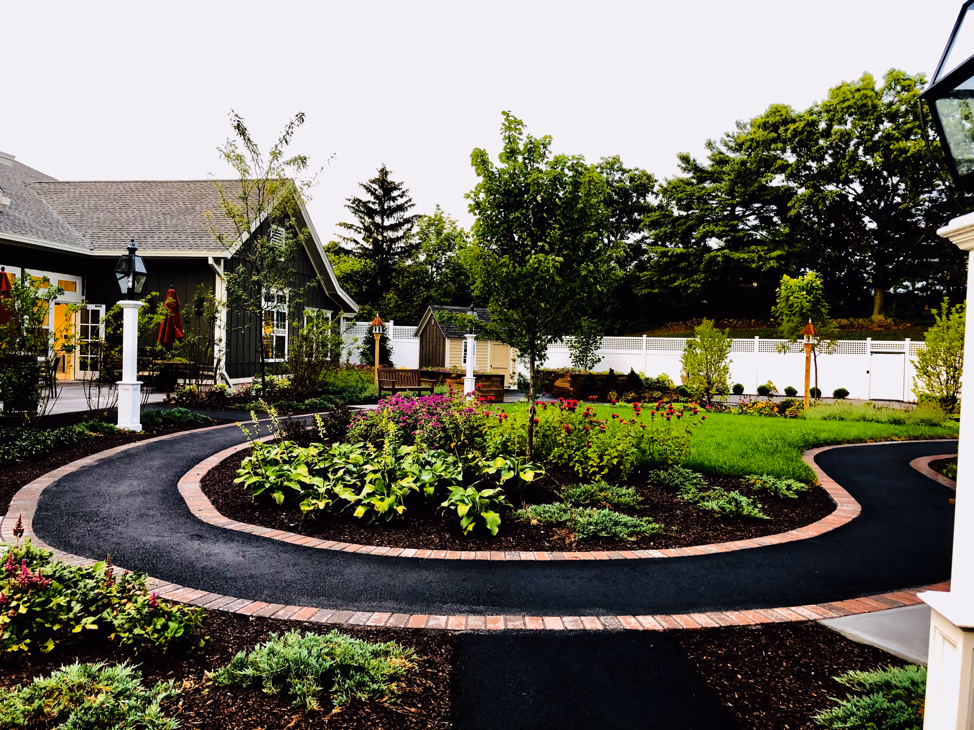 A landscaped outdoor garden area with a curved paved walkway bordered by bricks. The garden features various plants, flowers, and small trees. There is a building with a gray exterior and white trim on the left side, and a white fence in the background. Several lamp posts and benches are also visible in the garden.
