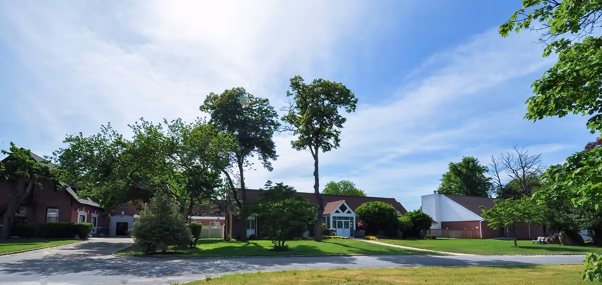 Exterior view of a senior living facility named Rosewood On the Sound, showing a single-story building with a peaked roof, surrounded by green trees and a well-maintained lawn under a partly cloudy blue sky.