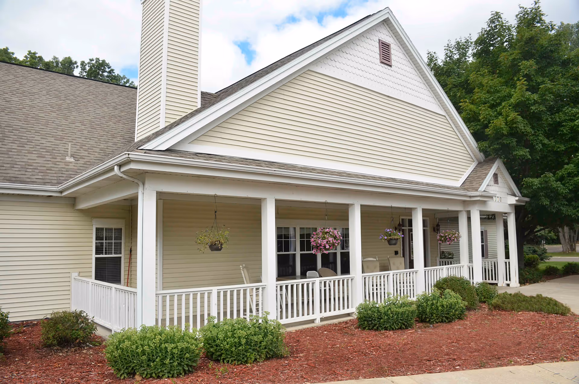 Exterior view of a beige senior living facility building with a covered porch featuring white railings and hanging flower pots. There are shrubs and mulch landscaping in front of the porch, and trees in the background under a partly cloudy sky.