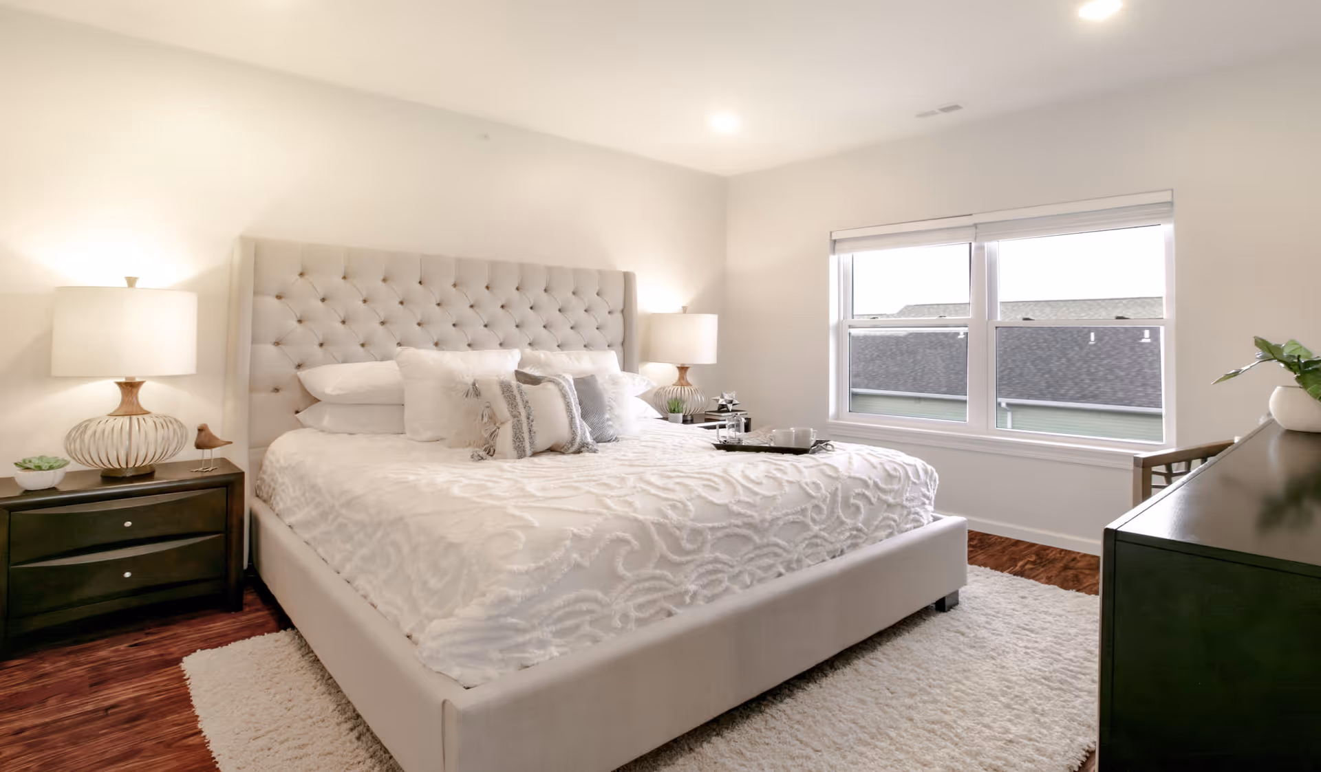 A bright and cozy bedroom with a large upholstered bed featuring a tufted headboard, white bedding, and decorative pillows. There are two bedside tables with matching lamps, a window with a view of rooftops, a dark dresser, and a light-colored rug on the wooden floor.
