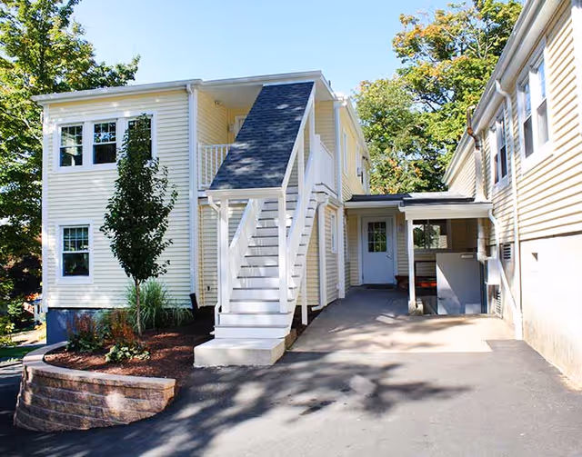 Exterior view of a light yellow two-story residential building with white trim. There is an outdoor staircase leading to the second floor, a small landscaped area with a tree and plants, and a paved driveway or walkway beside the building. The sky is clear and blue with some trees in the background.