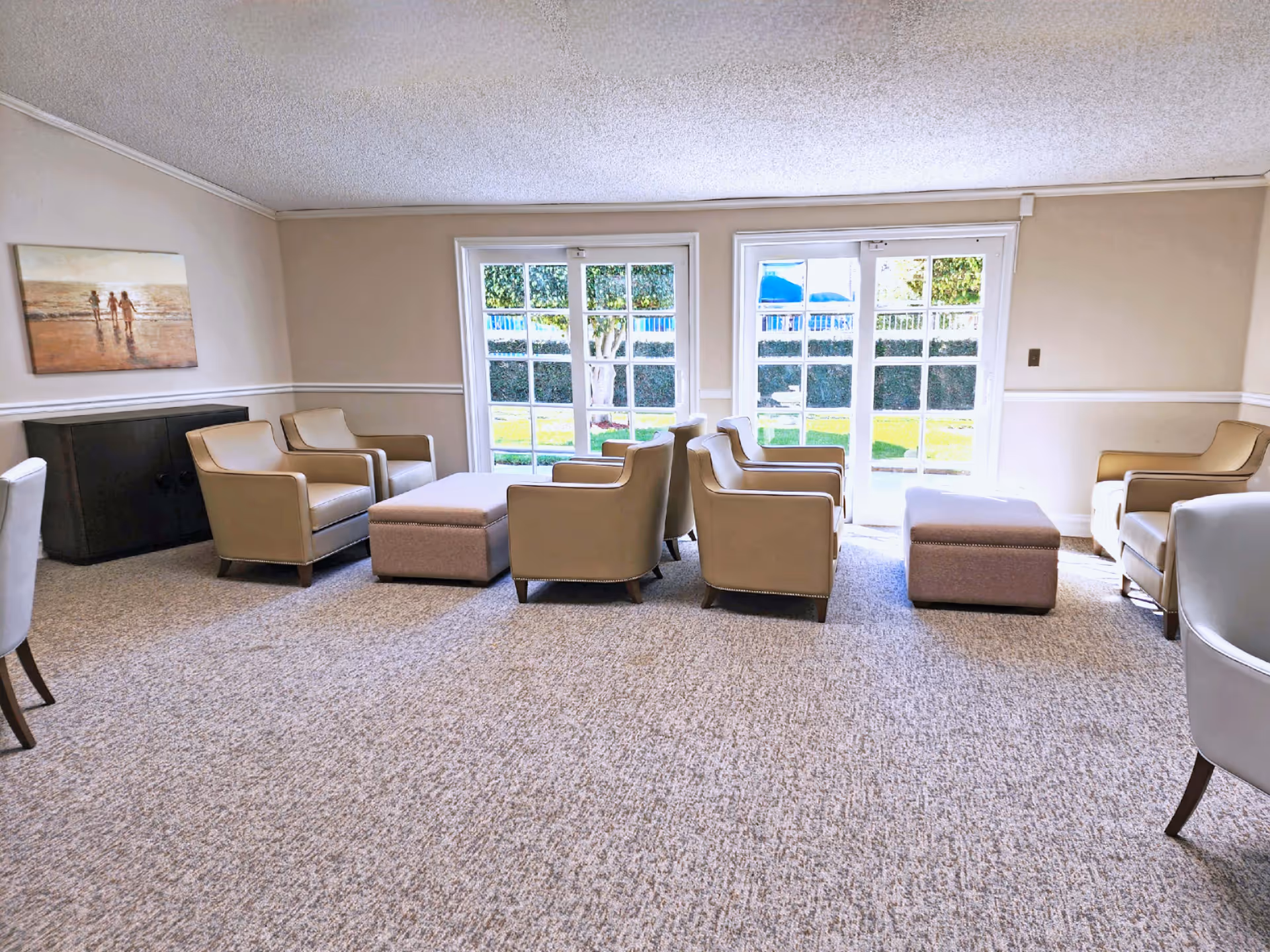 A bright and spacious living room area with beige walls and carpeted floor. The room features several beige armchairs arranged around two matching ottomans. Large glass doors and windows let in natural light and provide a view of a green outdoor space. A painting of three people walking on a beach hangs on the left wall above a dark cabinet.