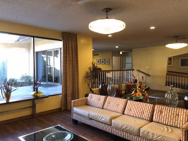 A cozy living room area with a long striped sofa adorned with multiple cushions. The room features large windows with curtains, allowing natural light to brighten the space. There are decorative vases with flowers on a glass coffee table and on a ledge by the window. The walls are painted yellow, and ceiling lights provide additional illumination. A railing and staircase are visible in the background.