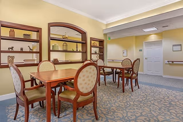 A well-lit room with two wooden tables and eight upholstered chairs arranged around them. The walls are painted light yellow, and there are built-in shelves with decorative items such as vases and small sculptures. The floor is carpeted with a patterned design, and there is a white door and handrails along the walls in the background.