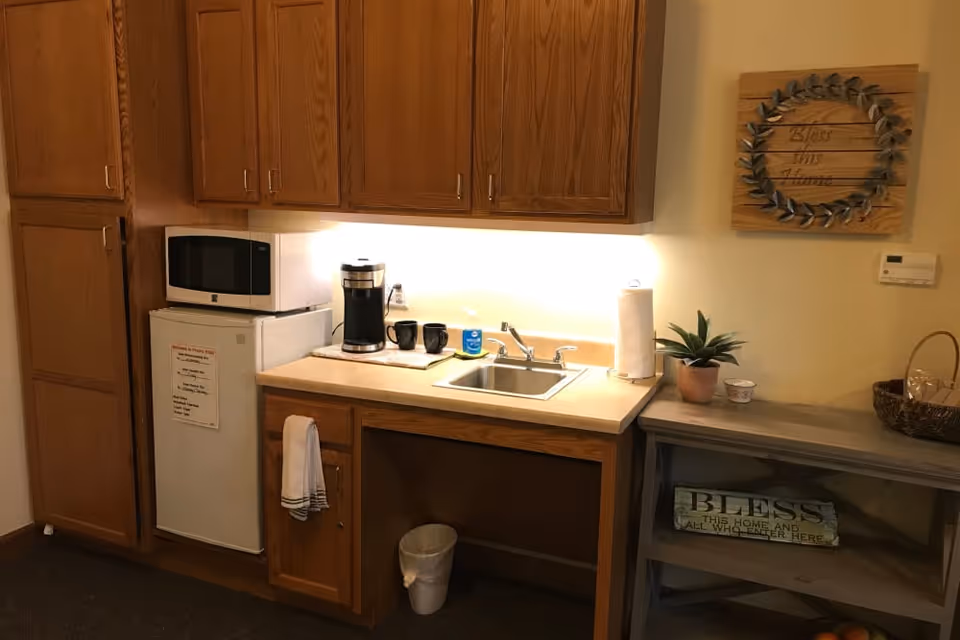 Small kitchenette area with wooden cabinets, a mini refrigerator with a microwave on top, a coffee maker with two black mugs, a sink with a paper towel holder, a small potted plant, and a decorative sign on the wall that reads 'Bless this Home'. Below the sink is a small trash can, and to the right is a gray shelf with a basket and a sign that says 'Bless this home and all who enter here'.